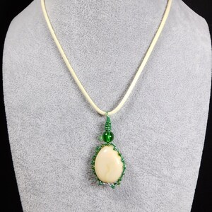 May include: A white oval pendant with a green wire wrap and green beads hangs from a cream-colored cord necklace.