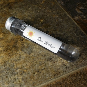 May include: A clear glass test tube with a silver lid and a white label that says "Sun Water". The label also features a yellow sun with orange rays.