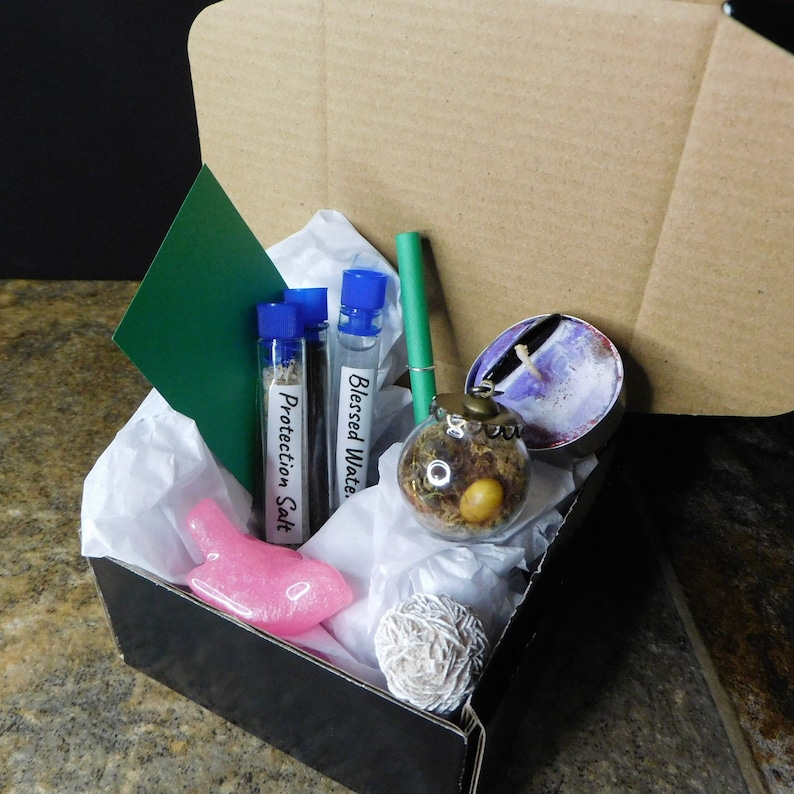 May include: A black box filled with various items, including a pink bird figurine, a small glass orb with dried flowers and a brown seed, a white candle, a green stick, and three small glass vials with blue caps labeled "Protection Salt", "Blessed Water", and "Blessed Water".