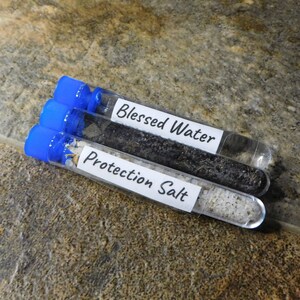 May include: Three small glass test tubes with blue screw-on caps. The tubes contain different substances: black sand, white salt, and clear liquid. The tubes are labeled "Blessed Water", "Protection Salt", and are on a brown speckled surface.