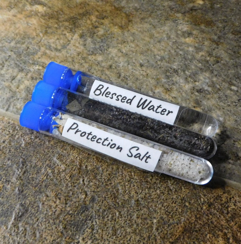 May include: Three small glass test tubes with blue screw-on caps. The tubes contain different substances: black sand, white salt, and clear liquid. The tubes are labeled "Blessed Water", "Protection Salt", and are on a brown speckled surface.