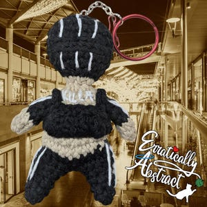 May include: A black and white crocheted doll with a black and white striped outfit and the word "KULLR" written on the chest. The doll is attached to a red and silver key chain.