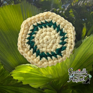 Puede incluir: Posavasos redondo de ganchillo color crema con un diseño geométrico verde oscuro. El posavasos tiene un aspecto texturizado. El fondo presenta hojas verdes grandes y el texto "Erraticly Abstract".