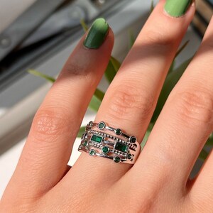 May include: A silver ring with multiple bands featuring emerald green stones. The ring is worn on a hand with green nail polish.