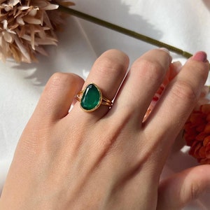 Peut inclure: Une bague en or avec une pierre précieuse verte en forme de larme. La bague est portée au doigt d'une personne.