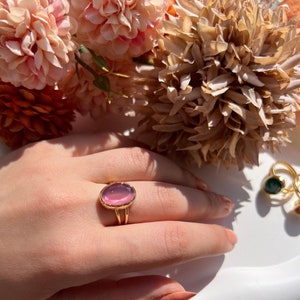 May include: A gold ring with a pink oval-shaped gemstone. The ring is on a finger and the background is blurred.