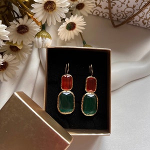 May include: A pair of gold-toned earrings with a rectangular, two-tone design. The top section is a reddish-orange color, while the bottom is a deep green. The earrings are displayed in a black box with a gold-colored exterior, next to white flowers.