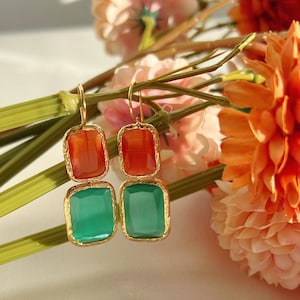 May include: A pair of gold-toned earrings with two rectangular stones. The top stone is orange and the bottom stone is green.