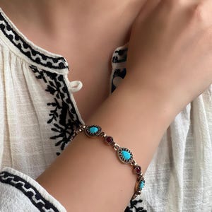 May include: A gold-toned bracelet featuring turquoise and red gemstones. The bracelet has an oval and teardrop design, with the gemstones set in a decorative metal setting. The bracelet is worn on the wrist.