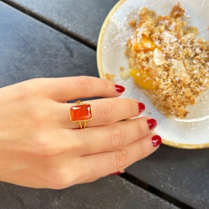Puede incluir: Un anillo de oro con una piedra roja rectangular. El anillo está en el dedo de una persona.