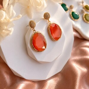 May include: A pair of drop earrings featuring faceted, oval-shaped orange stones set in gold-toned metal frames. Each earring has a textured, circular gold-toned stud top. The stones have a smooth, polished finish with a vibrant, translucent orange hue.
