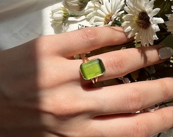 May include: A gold ring with a square green gemstone. The ring is on a finger.