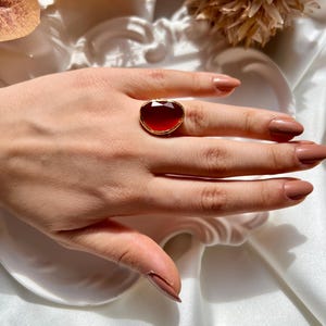 May include: A gold ring with a large, red, faceted gemstone. The ring is on a finger with light brown nail polish.