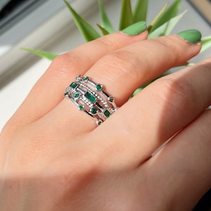 May include: A silver ring with multiple bands adorned with emerald green square and round gemstones.