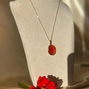 May include: A gold necklace with an oval, orange pendant. The necklace is displayed on a mannequin. A red flower is in the foreground. The pendant is approximately 2.5 cm in length.