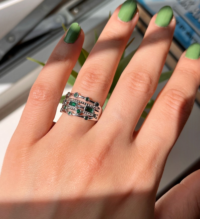 May include: A silver ring with multiple bands featuring emerald green stones. The ring is worn on a hand with green nail polish.