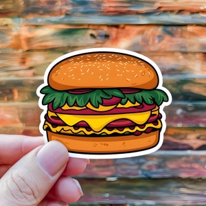 May include: A colorful sticker featuring a cartoon illustration of a double-decker burger. The burger has a sesame seed bun, lettuce, red onion, two cheese slices, and two meat patties. The sticker is held by a hand.