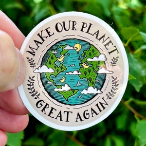 Könnte beinhalten: Ein runder Aufkleber mit beigefarbenem Hintergrund und einer grünen und blauen Illustration der Erde. Der Text "Make Our Planet Great Again" ist in brauner Schrift um die Erde geschrieben.