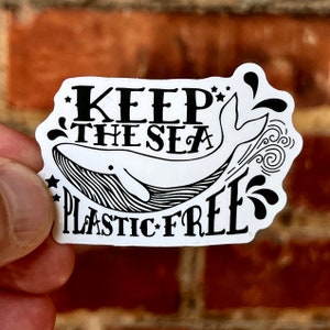 Puede incluir: Una pegatina blanca con una ilustración de ballena negra y el texto "Keep the Sea Plastic Free".