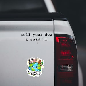 May include: A white vinyl sticker featuring the black text 'tell your dog i said hi' above a colorful illustration of a smiling Earth globe surrounded by a cow, pig, sheep, chicken, and duck, with the text 'equality for all' at the bottom.