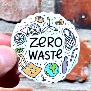 Puede incluir: Una pegatina blanca con un contorno negro y una variedad de ilustraciones que incluyen una bicicleta, una bombilla, una bolsa reutilizable, una botella de agua, una pajita, un globo terráqueo y una hoja. El texto "zero waste" está escrito en letras negras.