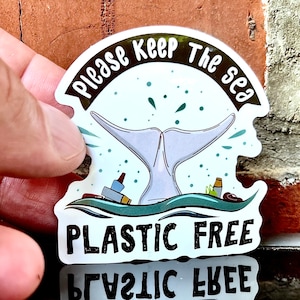 Puede incluir: Una pegatina con una cola de ballena blanca con el texto "Please Keep The Sea Plastic Free" en letras negras. La pegatina está en una pared de ladrillo.