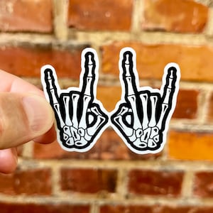 May include: Two black and white skeleton hands making the rock and roll hand gesture. The hands are on a brick background.