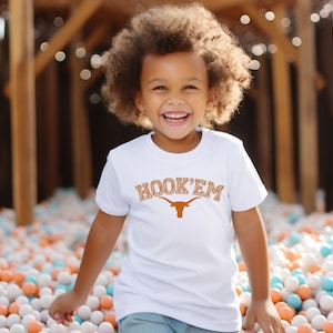 May include: A young child wearing a white t-shirt with an orange and brown "HOOK'EM" graphic and a Longhorn steer logo. The child is smiling and standing in a ball pit.
