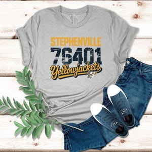 May include: A gray t-shirt with the text "Stephenville 76401 Yellowjackets" in yellow and navy blue. The shirt is paired with blue jeans and navy blue sneakers with white soles.
