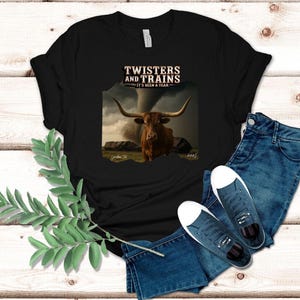 May include: Black t-shirt featuring a graphic of a longhorn steer with a tornado in the background. The text "Twisters and Trains" and "It's Been a Year" is printed above the image. The shirt is paired with blue jeans and sneakers.