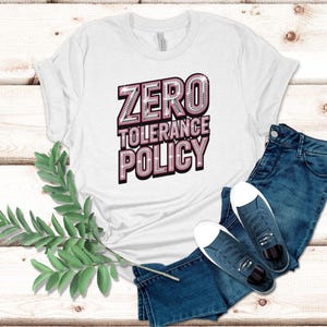 May include: White t-shirt with the text "ZERO TOLERANCE POLICY" in a pink and silver design. The shirt is paired with blue denim jeans and dark blue sneakers with white soles. A sprig of green leaves is on the left.