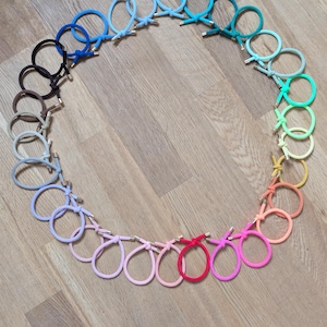 Könnte beinhalten: Eine kreisförmige Anordnung zahlreicher Haargummis in einem Farbspektrum aus Blau, Grün, Gelb, Orange, Pink, Lila, Grau, Braun und Schwarz. Jedes Haargummi ist mit einem kleinen, zylindrischen, goldfarbenen Metallelement versehen.