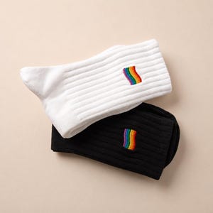 May include: A pair of ribbed crew socks, one white and one black, each featuring a small, embroidered rectangular patch with a vertical rainbow-coloured wavy stripe pattern centred on the upper portion of the fabric.