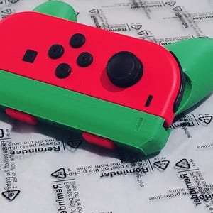 Könnte beinhalten: Ein roter und grüner Nintendo Switch-Controller mit Wassermelonen-Design. Der Controller hat einen schwarzen Joystick und vier schwarze Tasten.