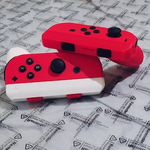 Puede incluir: Dos mandos de juego rojos y blancos. El mando superior es rojo con botones negros y un joystick. El mando inferior es rojo y blanco con botones negros y un joystick. Los mandos están sobre una superficie blanca con texto.