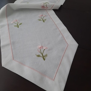 Op de afbeelding: Een witte tafelrunner met een roze geborduurd bloemenontwerp. De runner heeft een roze rand en is gemaakt van een zachte, linnenachtige stof.