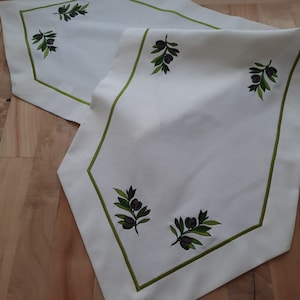 May include: White table runner with a green border and embroidered olive branches. The runner has a subtle sheen and is perfect for adding a touch of elegance to any table setting.