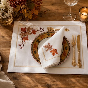 Op de afbeelding: Een herfsttafelsetting met een witte placemat met geborduurde herfstbladeren. Een bord met een bladerenpatroon, een gevouwen servet met bladborduurwerk en gouden bestek zijn op de placemat geplaatst. Een brandende kaars en een wijnglas zijn ook aanwezig.