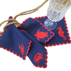 Puede incluir: Cuatro servilletas de lino azul con bordes festoneados rojos y criaturas marinas bordadas en rojo. Las servilletas presentan un caballito de mar, una tortuga, un cangrejo y una langosta.