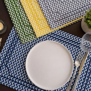 May include: A set of rectangular placemats featuring a chevron pattern in blue, green, yellow, and gray. Each placemat has a stitched border detail. A white plate, a silver fork, a silver knife, and a clear glass are placed on the blue mat.