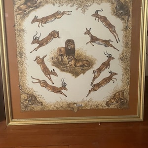 May include: A framed print of a vintage Hermes Paris scarf featuring a brown and white design of various animals, including a lion, a cheetah, and several gazelles, against a beige background. The text "Hermes Paris" and "Kenya" are visible in the print.