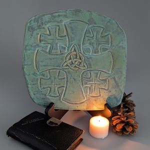 May include: A green ceramic plate with a Celtic knot design and four crosses carved into the surface. The plate is on a stand and there is a candle and a pine cone in the foreground.