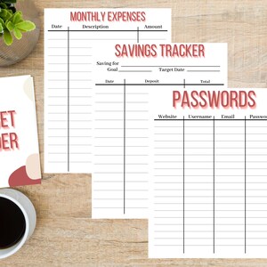 May include: A budget binder with a cover that reads "BUDGET BINDER" in red text. Inside are three white sheets for tracking monthly expenses, savings, and passwords. The sheets have black lines and headings in red. A cup of coffee and a plant are also visible.