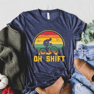 May include: A navy blue t-shirt with a vintage-style graphic of a person riding a bicycle in front of a sunset. The text "OH SHIFT" is printed below the graphic.
