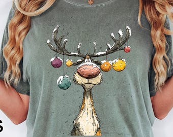 以下が含まれることがあります： モスグリーンのTシャツ。トナカイのイラストが特徴で、角にはカラフルなオーナメントが飾られています。左下には黒字で「Moss」の文字。柔らかく快適な素材で作られています。
