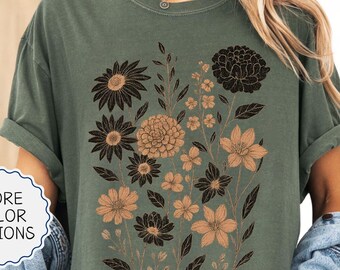 May include: Moss green t-shirt featuring a floral design in black and tan. The graphic includes various flower types and foliage. The shirt has rolled-up sleeves and a relaxed fit. The text "MORE COLOR OPTIONS" is in a white circle.
