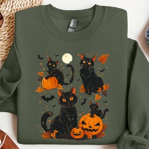 May include: A green sweatshirt with a Halloween-themed design featuring black cats and pumpkins. The design includes a full moon, bats, and autumn leaves.