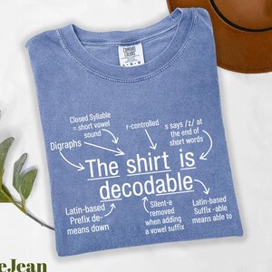 May include: A blue jean colored t-shirt with white text that reads The shirt is decodable, surrounded by linguistic annotations including Closed Syllable, r-controlled, s says /z/, Digraphs, Latin-based Prefix de-, Silent-e, and Latin-based Suffix -able.