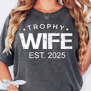 May include: A dark gray t-shirt with white text that reads "Trophy Wife Est. 2025".