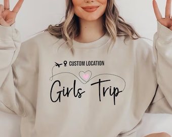 May include: A beige sweatshirt with a black graphic design that says "Girls Trip" with a pink heart and a plane icon. The text "Custom Location" is above the graphic.